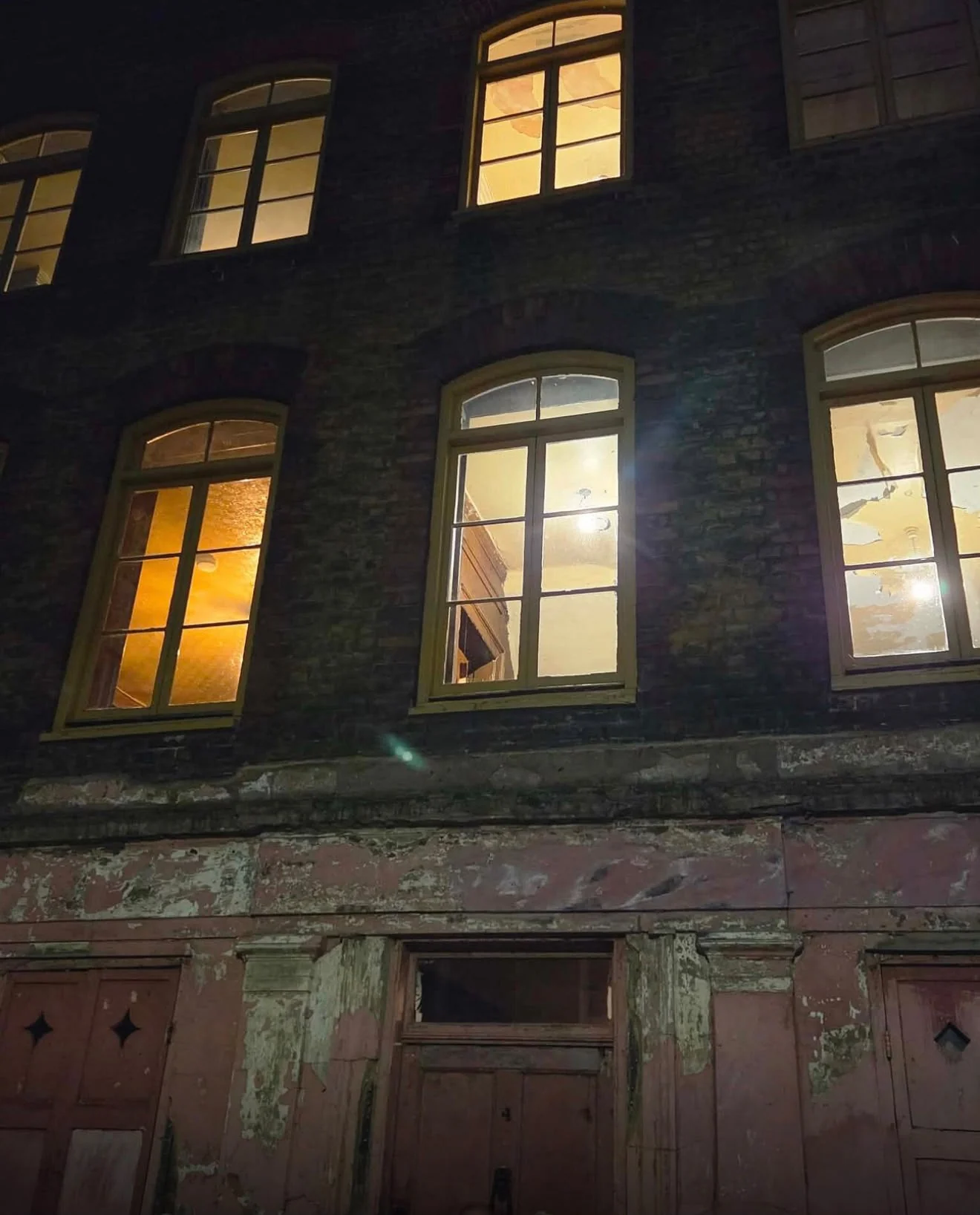 Low-angle nighttime view of a weathered brick building with glowing arched windows, peeling paint and old wooden doors.